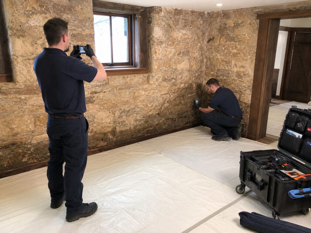Two technicians photograph the water line and record moisture readings in an Alton stone basement.